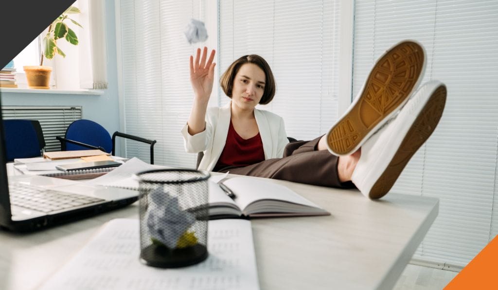 Woman sitting at an office with her feet on the desk tossing paper in a basket on the desk thinking how to combat quiet quitting